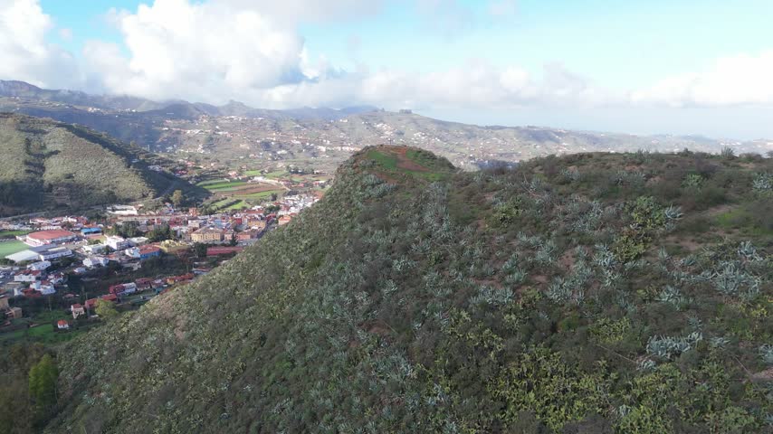 Aerial drone shot of a charming town nestled in a lush valley on Gran Canaria. The camera pushes in, revealing Mirador de Conesa with whitewashed houses and cultivated fields below.