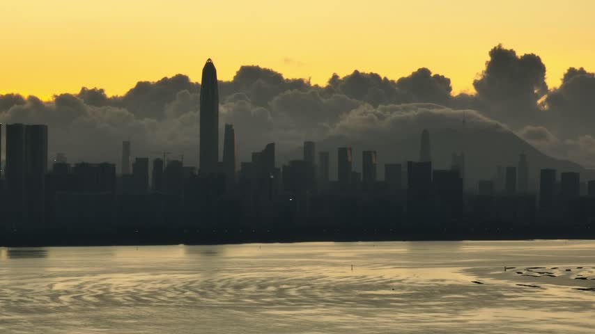 Elevated view of dark waterfront skyline above rippled golden water in Shenzhen, Guangdong, China. The scene features Ping An Finance Centre. Relevant to architecture and urban planning.