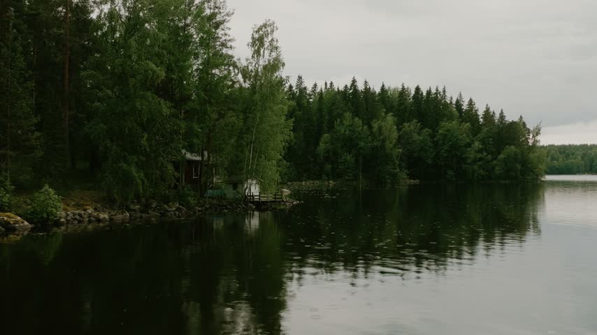 Aerial view of a small red cabin nestled on the forested lakeshore under an overcast sky in Hameenlinna, Kanta-Hame, Finland.