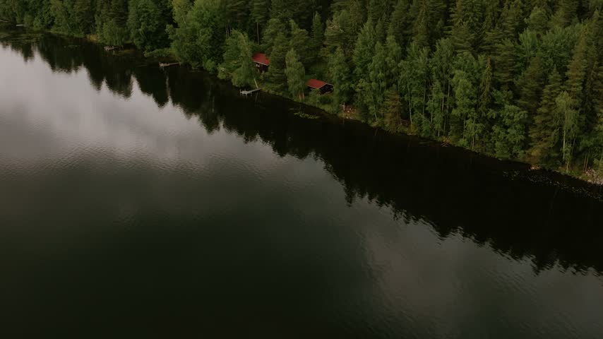 Aerial view of a calm lake surrounded by dense pine forest with a small red cabin on the shore in Hameenlinna, Kanta-Hame, Finland.