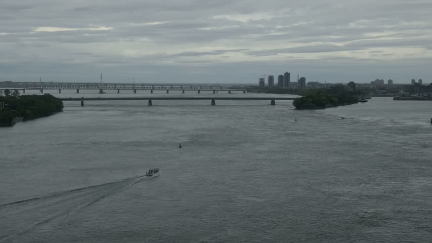 St. Lawrence river near Jacques Cartier bridge, Montreal, with small boat leaving white wake, distant bridges and city towers under grey cloudy sky, Quebec, Canada.