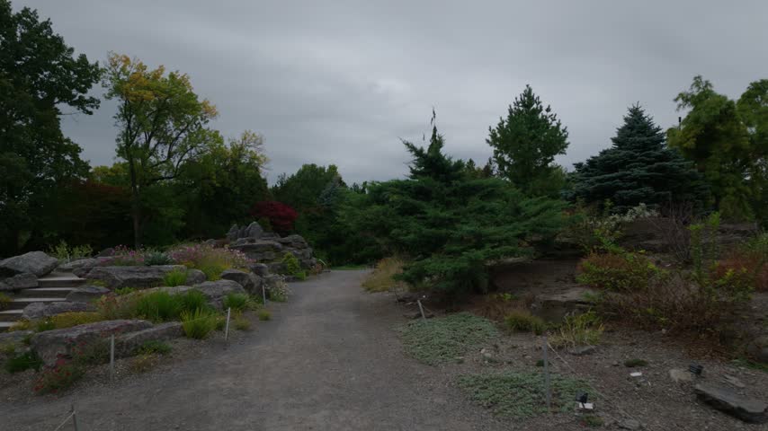 Montreal Botanical garden gravel path through rock garden with evergreens, colorful autumn trees and flowering plants, walk in shot under grey sky, Quebec, Canada.