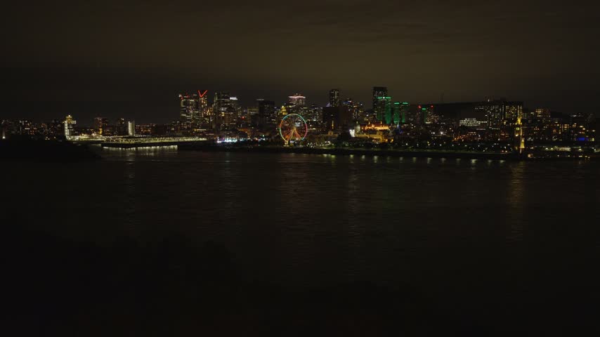 Montreal skyline and Old Port Ferris wheel lit at night across St. Lawrence river, drone pushes forward from Parc Jean-Drapeau, calm dark water reflections, Quebec, Canada.