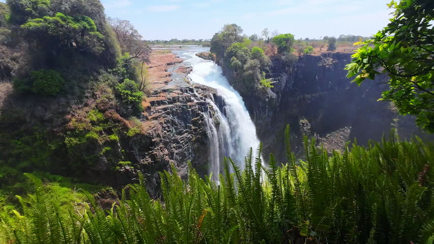 Water plunges over exposed rock into a steep gorge at Victoria Falls during the dry season, framed by green vegetation.