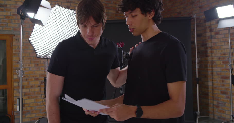 Diverse male colleagues in studio wearing black tees, placing guiding hand and rehearsing lines. Collaboration, script, dialogue, director, coaching, brick, backdrop