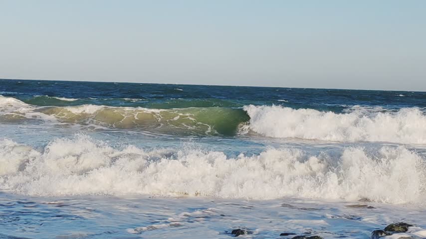  Powerful ocean waves crashing forcefully against the shoreline, creating a dramatic scene of natural beauty and untamed energy. The roaring waves rise high above the sea surface before rushing toward the beach, forming thick layers of white foam and sparkling sea spray. As the water strikes the shore, it spreads rapidly across the wet sand and then retreats into the ocean, leaving behind swirling patterns of foam. The stormy sea appears rough and restless, with waves continuously building, breaking, and colliding against one another. The deep blue and grey tones of the water create a striking contrast with the bright white froth along the wave crests. The powerful movement of the ocean highlights the immense strength of nature and the endless rhythm of the tides.