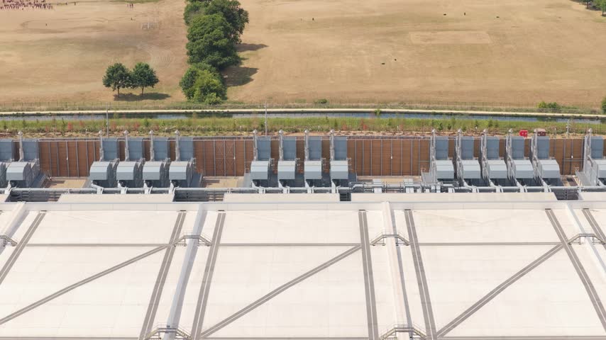 A wide overhead shot shows the exterior of a modern data center with many cooling units installed along the edge of a white flat roof near a grassy field and trees.