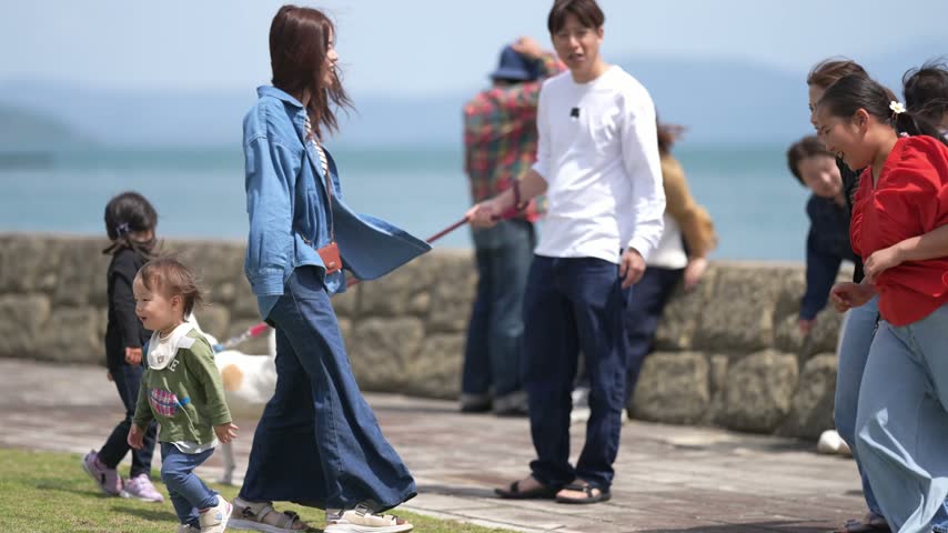 Bright day near sea with people and dogs. Woman in blue walks with white dog on leash. Children and adults enjoy sunny seaside path. Dogs and humans interact happily, casually