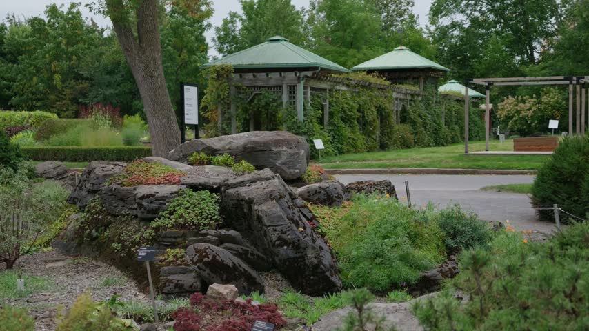 Montreal Botanical garden rock garden with shrubs swaying in wind, green roof pavilions, vine covered trellis, lawn and path on overcast day, Quebec, Canada.