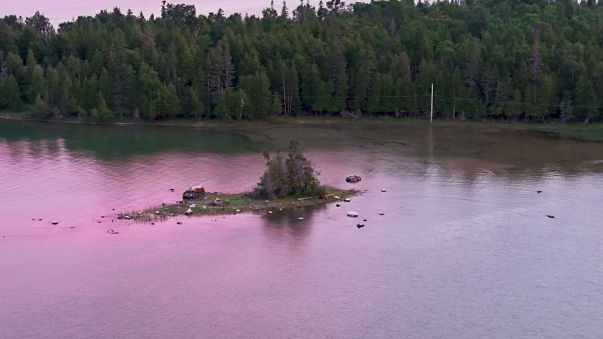 Aerial view of a small rocky island with shrubs surrounded by pink twilight reflections and a forested shoreline near Hessel Michigan
