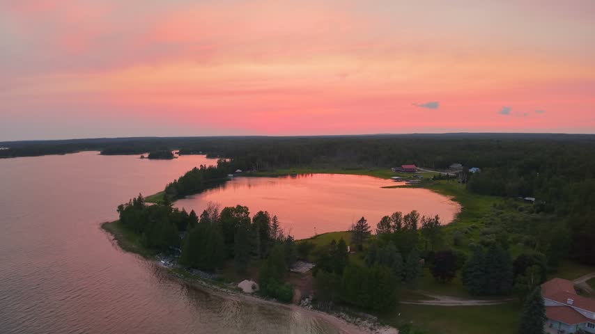 Drone panorama of a wooded shoreline curving around an inlet beneath an orange and pink sunset sky near Hessel Michigan