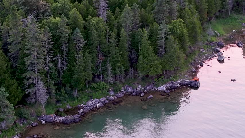 Aerial view of dense woodland and a rocky shoreline beside calm reflective water near Hessel in the Upper Peninsula of Michigan