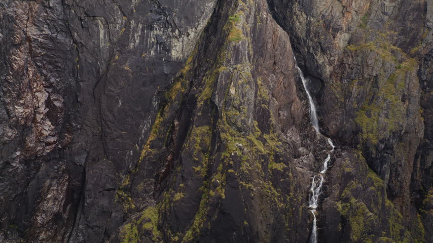 Thin waterfall runs down a sheer mossy cliff wall, framed tight on wet stone textures. Atmospheric cutaway for geology, wilderness, or adventure travel projects.