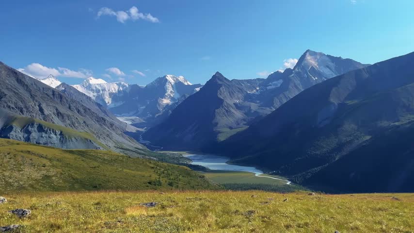 Sweeping mountain valley displaying turquoise glacial lake nestled between towering snow capped peaks, green alpine meadow in foreground, clear blue sky.