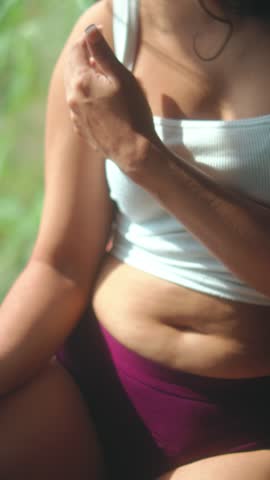 Woman gently touches her upper arm while seated in warm sunlight, wearing a pale crop top. A sensual close detail for body confidence, beauty and relaxed lifestyle content.