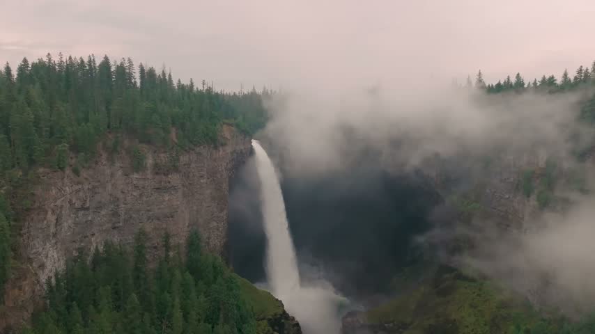 Aerial drone footage of the towering Helmcken Falls plunging into a misty forested gorge with cloud rolling over the cliffs in Wells Gray Park British Columbia Canada