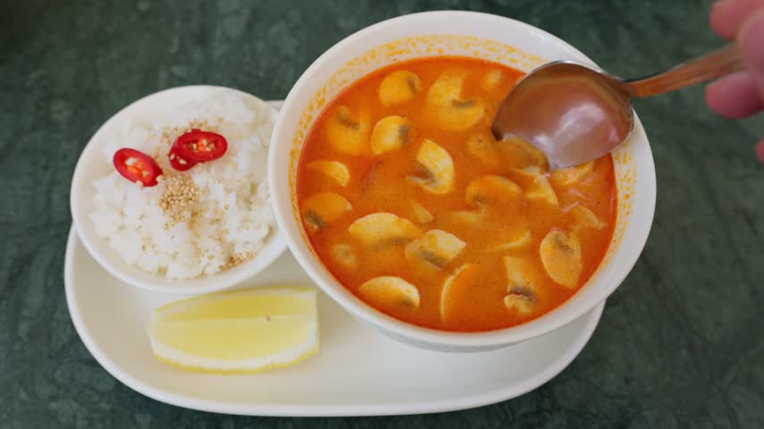 Close up handheld shot of spoon lifting spicy shrimp soup with mushrooms and tomatoes near bowl of steamed jasmine rice.