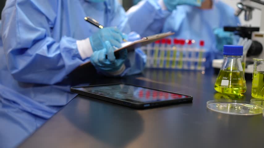 Scientists in protective suits and gloves use a tablet beside lab glassware with yellow liquid, showing modern medical research and testing.