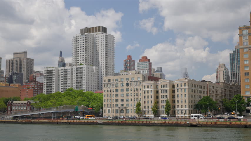 Apartment towers and prewar buildings line FDR Drive on the East River in New York City, New York, USA. Pedestrian bridge, green trees, yellow taxis and traffic under sunny summer clouds.