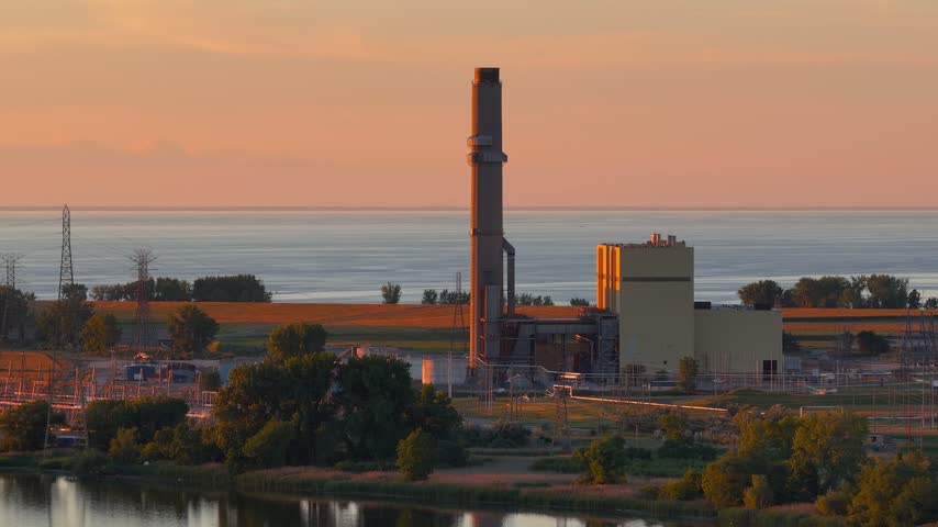 Industrial landscape showing a power station with a tall smokestack near the water at sunset.