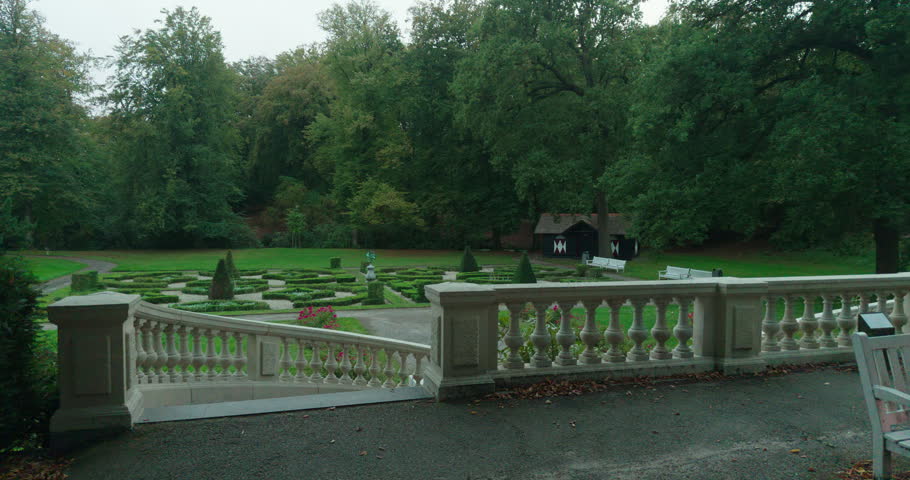 A formal garden in The Hague, Netherlands, Europe, with low hedges, gravel pathways, pink blossoms and conical topiary trees. Fallen leaves lie along a curved stone balustrade.