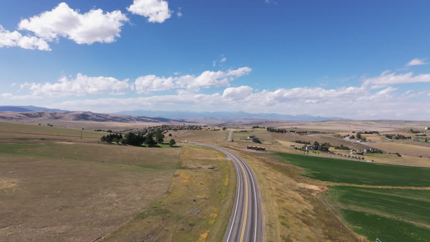 Aerial view of a rural Montana highway winding through open farmland, rolling hills, distant mountains, scattered homes, and bright summer skies, with vehicles traveling ahead.