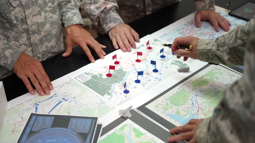 Military team planning a field operation around detailed maps with colored markers, navigation tools, and digital displays on a briefing table