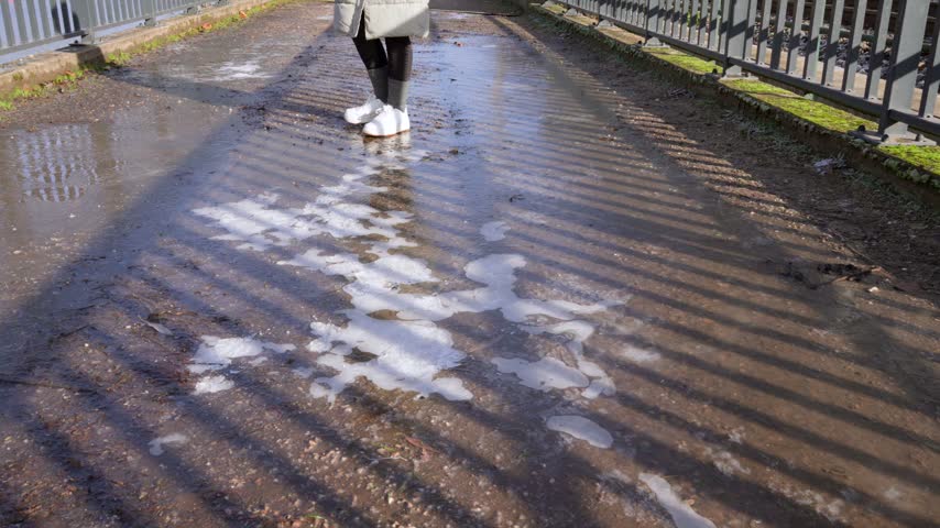 A person walks along a wet winter path lined with snow and fence shadows.