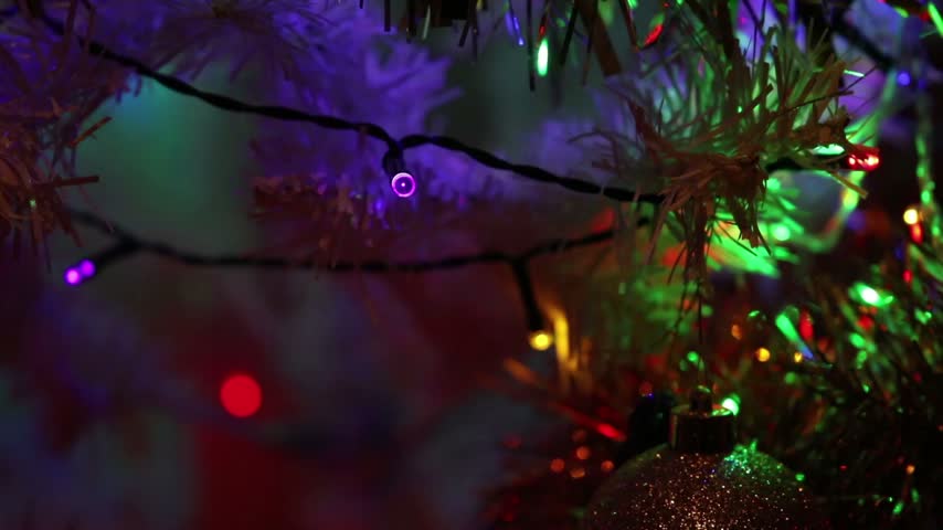 A close-up view of a white Christmas tree decorated with multicolored LED lights gently twinkling, shimmering tinsel, and Christmas ornaments.