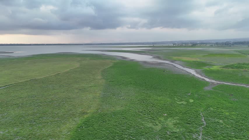 Aerial panorama of lush green fields and a winding stream connecting to a wide reservoir under a dramatic cloudy sky, depicting natural outdoor beauty.