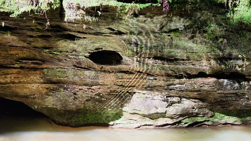 Slow pan across a shaded sandstone overhang as rippling creek light dances on the stone. A dark cave mouth sits at the waterline. Daytime, no people. Old Man