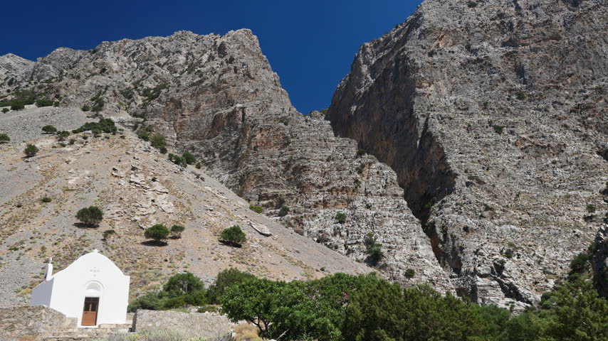 Small white whitewashed chapel standing at entrance of steep rocky gorge, Ha Gorge in Crete, Greece, wide establishing shot
