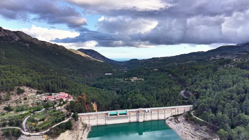 Forest Valley Dam Seen From Hillside With Dense Pine Canopy, Winding Road And Distant Village Beneath Heavy