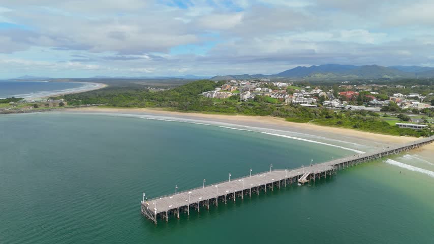Aerial drone tracking shot alongside the historic timber jetty and sandy beach at Coffs Harbour, New South Wales, Australia, with mountain ranges