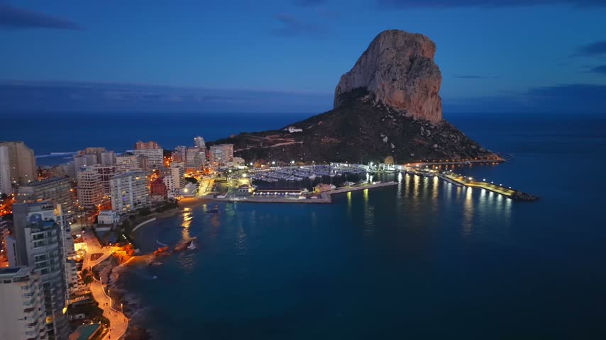 Dusk Aerial View Of Illuminated Marina Towering Limestone Rock Overlooks Calm Bay City Lights Reflecting