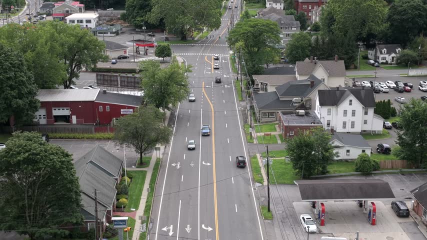 A quiet small town intersection in New York state spreads below, a red barn like building, white clapboard houses and a filling station bordering tree lined streets. Aerial approaching shot