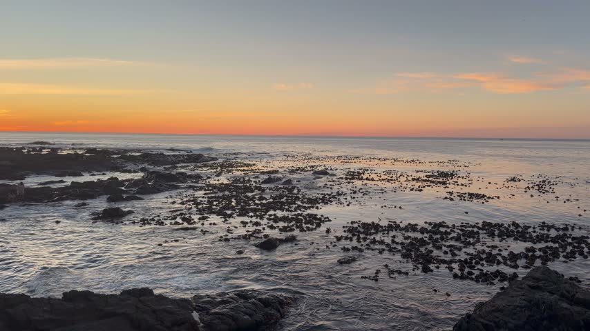 A vivid orange and peach sunset glows above a calm Atlantic horizon, while kelp and dark rocks are scattered across shallow tidal water at Sea Point in Cape Town, South Africa.