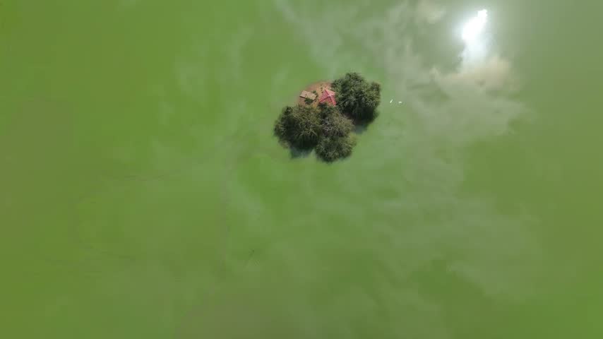 An overhead aerial view looking straight down at a small island with a red-roofed structure and trees in a green lake in Baalbek, Lebanon.