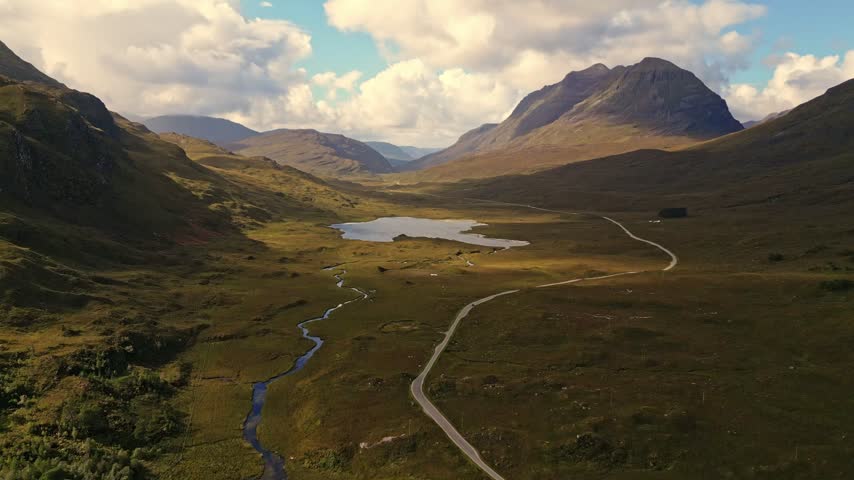 Aerial view of winding road, stream, and mountains under dramatic clouds in rural Scotland