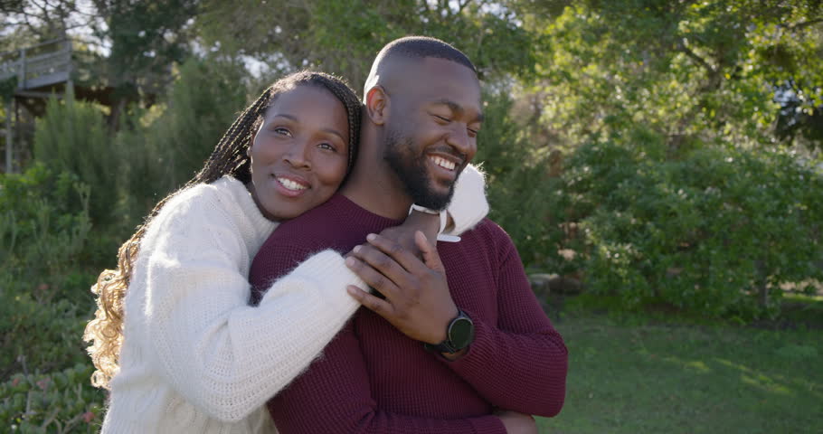 Romantic couple woman approaching, wrapping arms around man holding phone in knits, posing on lawn. Outdoor, garden, greenery, embrace, affection, togetherness, portrait