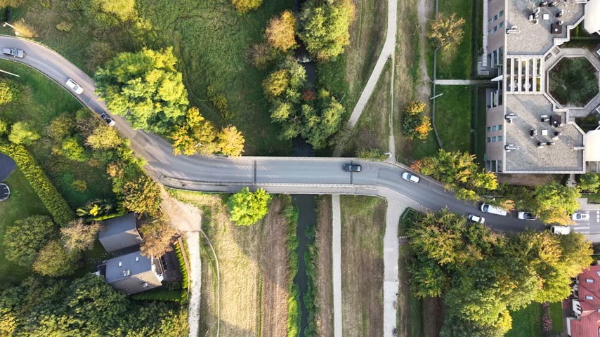 Aerial view of cars crossing a narrow bridge over a small stream beside rural homes and autumn trees in a green countryside landscape. Traffic at country landscape surrounded by trees, grassy fields, paths, and homes