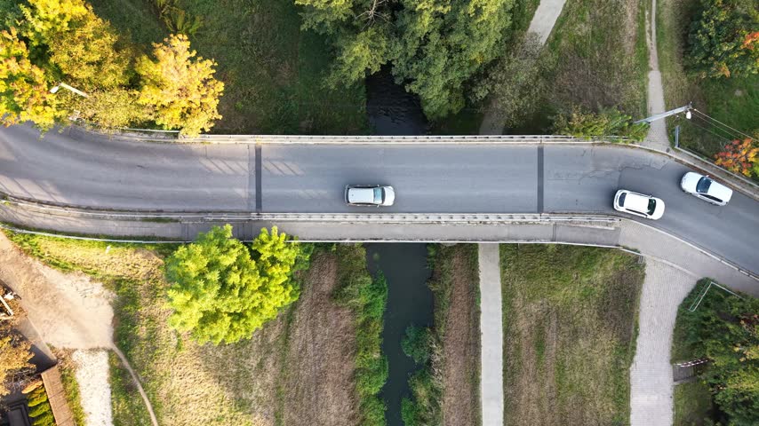 Aerial view of cars crossing a narrow bridge over a small stream beside rural homes and autumn trees in a green countryside landscape. Traffic at country landscape surrounded by trees, grassy fields, paths, and homes