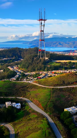 Tower on forested hill above winding road, San Francisco. Red and white radio mast, green grassy slopes, houses on the ridge, curving asphalt road and bay in distance. Vertical video.