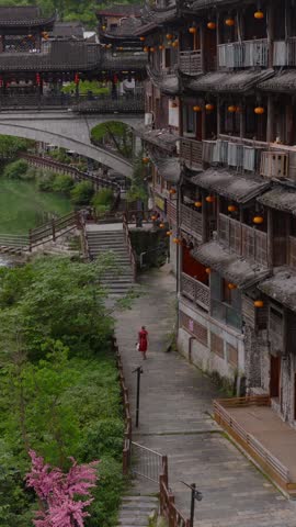 Telephoto pull back along old wooden stilt houses following a woman in a red dress on the stone path, covered bridge behind, Furong ancient town, Hunan, China