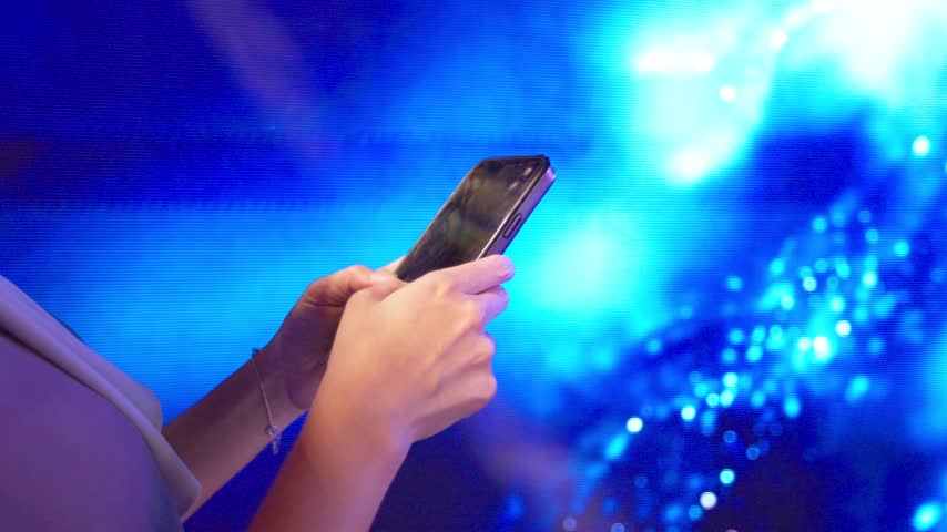 Hands hold and tap a black smartphone in front of a blue digital display, with luminous circles and flowing light patterns behind the device in a close cropped view of fingers using the touchscreen.
