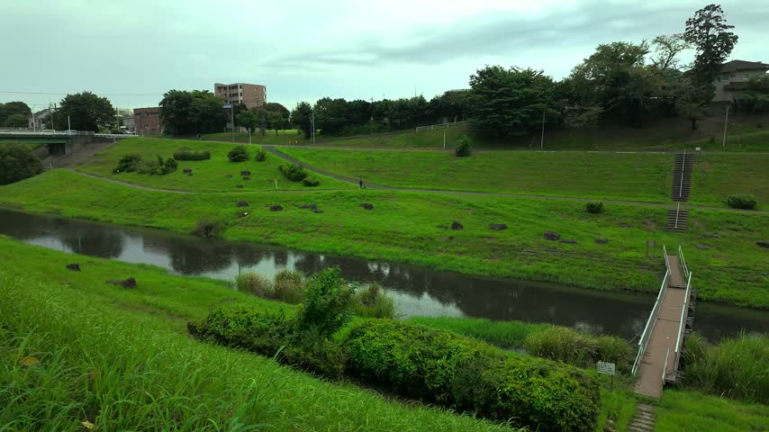 Rainy day at a lush green riverside park in Japan, small bridge over a stream, people walking with umbrellas on the bank.