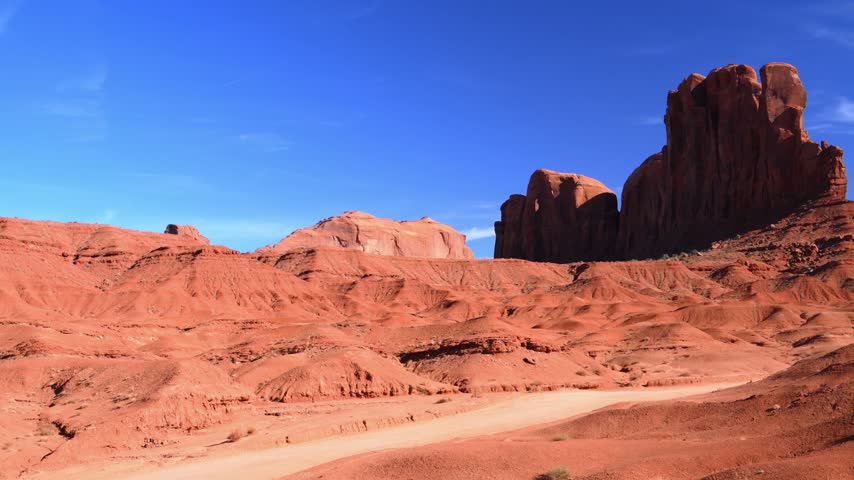 A majestic desert landscape of Monument Valley, featuring vivid reddish-orange sandstone buttes against a bright blue sky. The main subject of the photograph is the famous Camel Butte, a sandstone rock formation whose outline resembles the silhouette of a reclining camel. A dirt road running along a popular tourist route is visible at the base of these ancient geological formations.