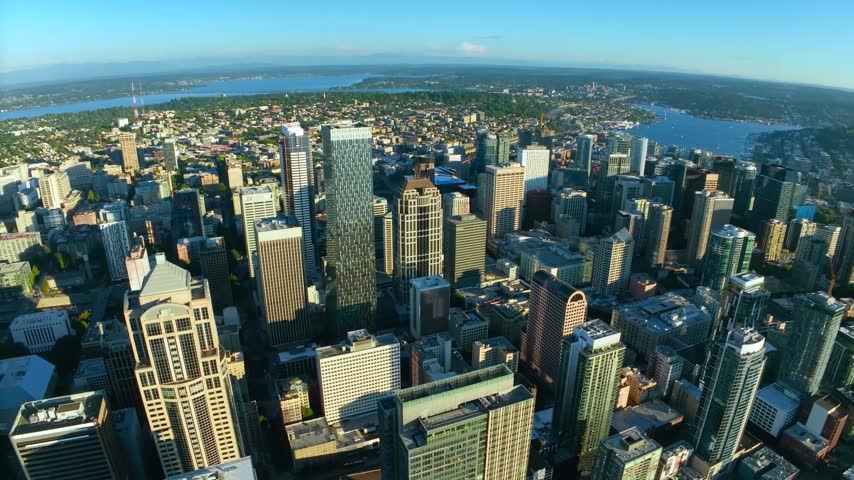 Wide high-altitude aerial drone perspective looking down at the dense corporate skyscrapers and office towers of the central business district in Seattle, Washington, highlighting the distinctive architecture of 1201 Third Avenue, the shadow textures of street canyons, and Lake Union visible in the background under a clear blue sky.