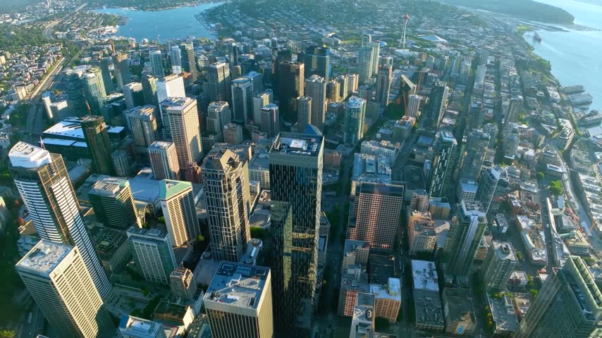 Wide fish-eye high-altitude aerial perspective looking north across the dense skyscraper grid of the central business district in Seattle, Washington, showcasing The Space Needle, the calm waters of Elliott Bay on the right, Lake Union on the left, and distant suburban peninsula horizons under a clear blue sky.