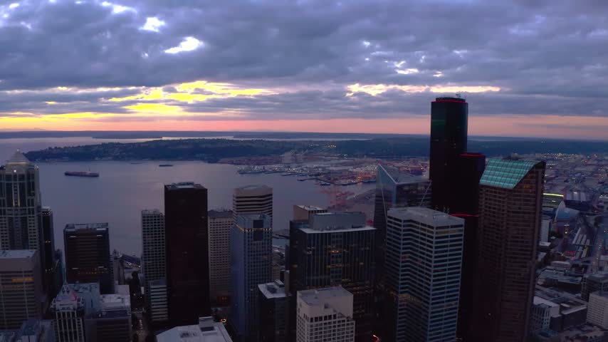 High-angle panoramic aerial drone perspective looking southwest over the corporate skyscrapers of downtown Seattle, Washington, showcasing Columbia Center, Smith Tower, and 1201 Third Avenue alongside the calm waters of Elliott Bay under a dramatic sunset twilight cloud deck.